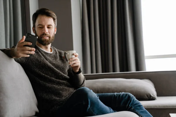 Bearded Man Using Mobile Phone Drinking Coffee While Sitting Sofa — Stock Photo, Image