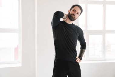 Sakin, orta yaşlı, sakallı, spor kıyafetli bir adam. Pencere kenarında durup, boynunu ovalıyor.