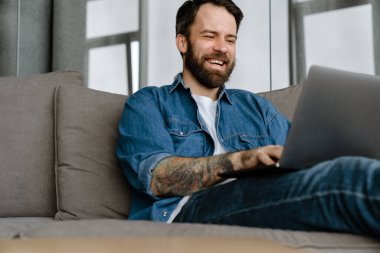 Sakallı Avrupalı adam evdeki kanepede otururken dizüstü bilgisayarla çalışıyor.