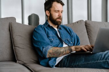 Sakallı Avrupalı adam evdeki kanepede otururken dizüstü bilgisayarla çalışıyor.