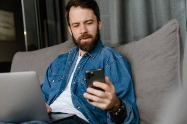 Sakallı adam evde kanepede otururken dizüstü bilgisayar ve cep telefonuyla çalışıyor.