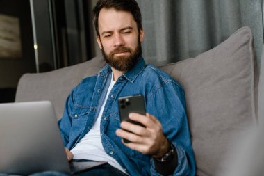 Sakallı adam evde kanepede otururken dizüstü bilgisayar ve cep telefonuyla çalışıyor.