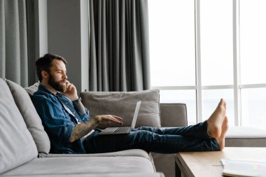 Evde kanepede dizüstü bilgisayarla çalışırken cep telefonuyla konuşan sakallı adam.