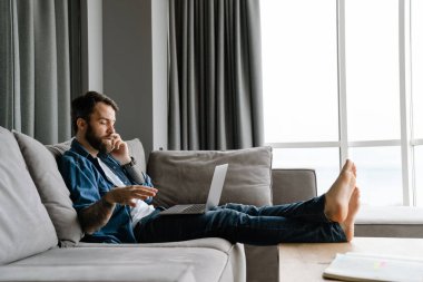 Evde kanepede dizüstü bilgisayarla çalışırken cep telefonuyla konuşan sakallı adam.
