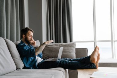 Evde kanepede dizüstü bilgisayarla çalışırken cep telefonuyla konuşan sakallı adam.