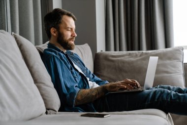 Sakallı Avrupalı adam evdeki kanepede otururken dizüstü bilgisayarla çalışıyor.