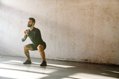 Orta yaşlı beyaz bir sporcunun gri duvarın içinde çömelmiş egzersiz yapması.