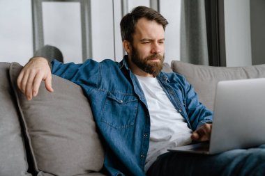 Sakallı Avrupalı adam evdeki kanepede otururken dizüstü bilgisayarla çalışıyor.