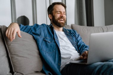 Sakallı adam evde kanepede otururken bilgisayarla gülüp çalışıyor.