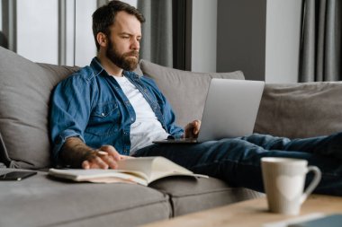 Sakallı adam evdeki kanepede dizüstü bilgisayarla çalışırken notlar yazıyor.