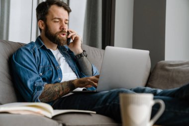 Sakallı adam evde çalışırken cep telefonuyla konuşuyor ve laptop kullanıyor.