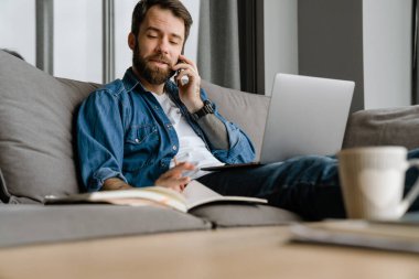 Sakallı adam evde çalışırken cep telefonuyla konuşuyor ve laptop kullanıyor.