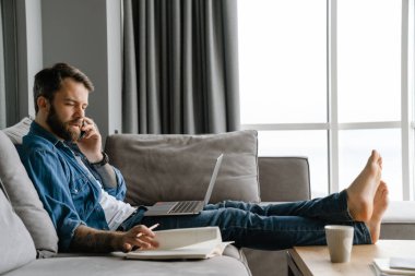 Sakallı adam evde çalışırken cep telefonuyla konuşuyor ve laptop kullanıyor.