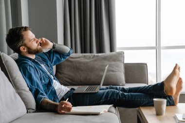 Sakallı adam evde çalışırken cep telefonuyla konuşuyor ve laptop kullanıyor.