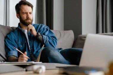 Sakallı Avrupalı adam evdeki kanepede otururken notlar yazıyor.