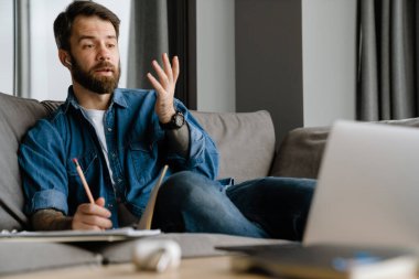 Sakallı Avrupalı adam evdeki kanepede otururken dizüstü bilgisayarla hareket ediyor ve çalışıyor.