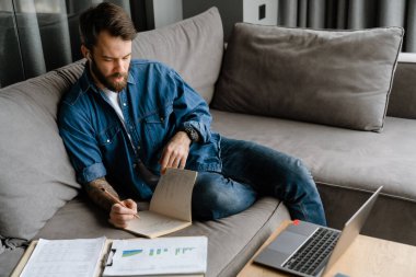 Sakallı Avrupalı adam evdeki kanepede otururken notlar yazıyor.