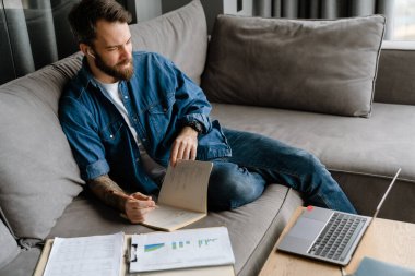 Sakallı adam evdeki kanepede dizüstü bilgisayarla çalışırken notlar yazıyor.