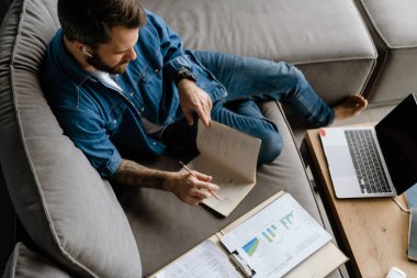 Sakallı adam evdeki kanepede dizüstü bilgisayarla çalışırken notlar yazıyor.