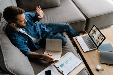 Sakallı adam evde dizüstü bilgisayarla konferans görüşmesi yaparken notlar yazıyor.