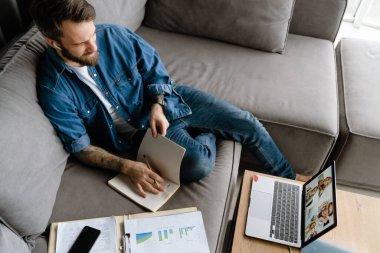 Sakallı adam evde dizüstü bilgisayarla konferans görüşmesi yaparken notlar yazıyor.