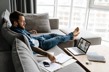 Sakallı adam evdeki kanepede otururken bilgisayarla konferans görüşmesi yapıyor.