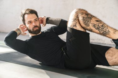 Genç, kendine güvenen esmer, sakallı sporcu içeride spor yaparken karın kasları çalışıyor.
