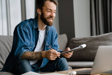 Sakallı Avrupalı adam evdeki kanepede otururken dizüstü bilgisayarla çalışıyor.