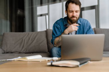 Sakallı Avrupalı adam evdeki kanepede otururken dizüstü bilgisayarla çalışıyor.