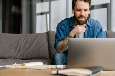 Sakallı Avrupalı adam evdeki kanepede otururken dizüstü bilgisayarla çalışıyor.