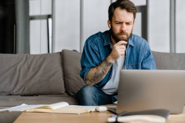 Sakallı Avrupalı adam evdeki kanepede otururken dizüstü bilgisayarla çalışıyor.