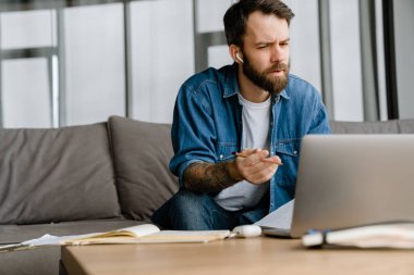 Sakallı Avrupalı adam evdeki kanepede otururken dizüstü bilgisayarla çalışıyor.