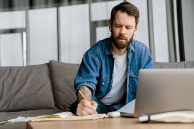 Sakallı adam evde dizüstü bilgisayarla konferans görüşmesi yaparken notlar yazıyor.