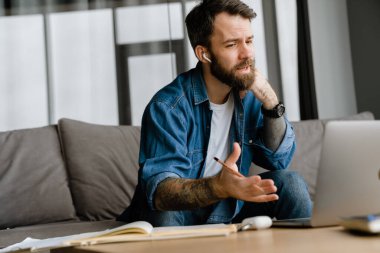 Sakallı Avrupalı adam evdeki kanepede otururken dizüstü bilgisayarla hareket ediyor ve çalışıyor.