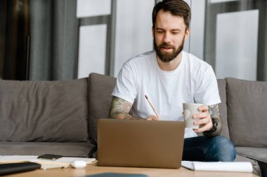 Evde dizüstü bilgisayarla çalışırken kahve içen sakallı Avrupalı adam.