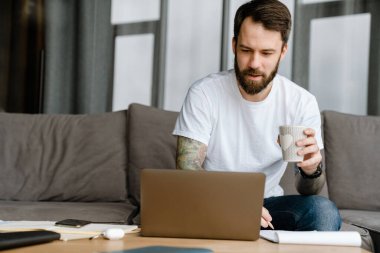 Evde dizüstü bilgisayarla çalışırken kahve içen sakallı Avrupalı adam.