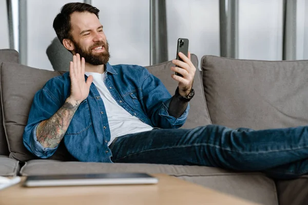 Sakallı Avrupalı adam evdeki kanepede otururken el kol hareketi yapıyor ve cep telefonu kullanıyor.