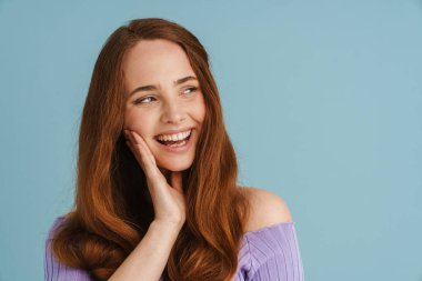 Mavi duvarın arkasında duran uzun kızıl saçlı, gülümseyen, sıradan bir kadının portresi.