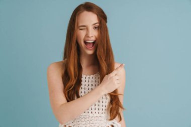 Genç kızıl, göz kırpan ve işaret eden heyecanlı kadın mavi arka planda izole edilmiş.