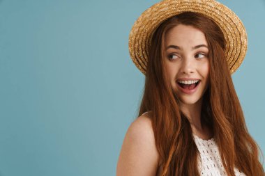 Hasır şapkalı genç kızıl kadın, gülümsüyor ve mavi arka planda tek başına bir kenara bakıyor.