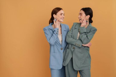 İki gülümseyen zarif kadın şık ceketler giyip birbirlerine yakın poz veriyorlar. Bej arka planda soyutlanmışlar.