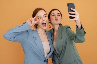 Sarı arka planda izole edilmiş cep telefonuyla selfie çeken iki genç kadın.