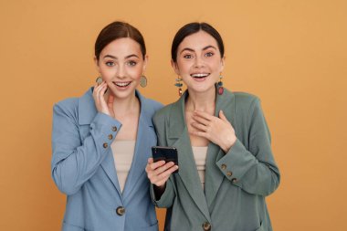 Sarı arka planda izole edilmiş cep telefonu kullanırken gülümseyen iki genç kadın.