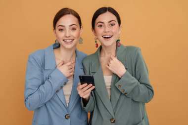 Sarı arka planda izole edilmiş cep telefonu kullanırken gülümseyen iki genç kadın.