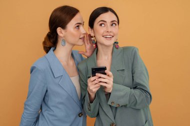 Sarı arka planda izole edilmiş cep telefonu kullanırken fısıldayan iki genç kadın.