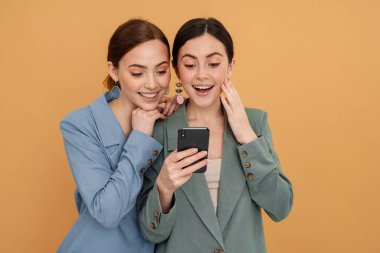 Sarı arka planda izole edilmiş cep telefonu kullanırken gülümseyen iki genç kadın.