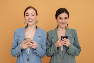 Sarı arka planda izole edilmiş cep telefonu kullanırken gülümseyen iki genç kadın.