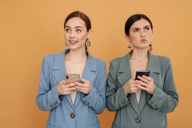 İki genç kadın cep telefonu kullanıyor ve sarı arka planda izole edilmiş farklı taraflara bakıyor.