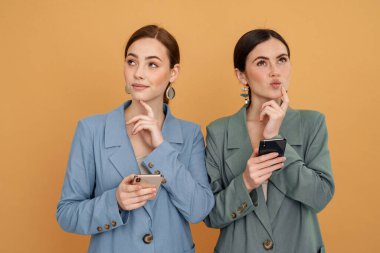 İki genç kadın cep telefonu kullanıyor ve sarı arka planda izole edilmiş farklı taraflara bakıyor.
