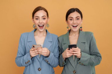 Sarı arka planda izole edilmiş cep telefonu kullanırken gülümseyen iki genç kadın.